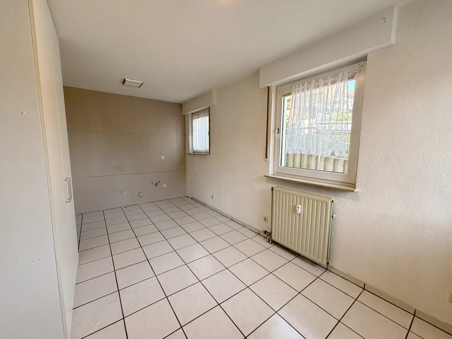 Empty room with white tiled floors, white walls, a radiator under a window with sheer curtains, and a built-in wardrobe.