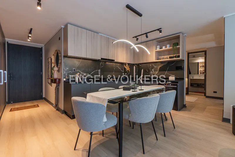 A modern apartment interior with a dining table, gray chairs, and a black marble kitchen. Overhead lighting illuminates the space.