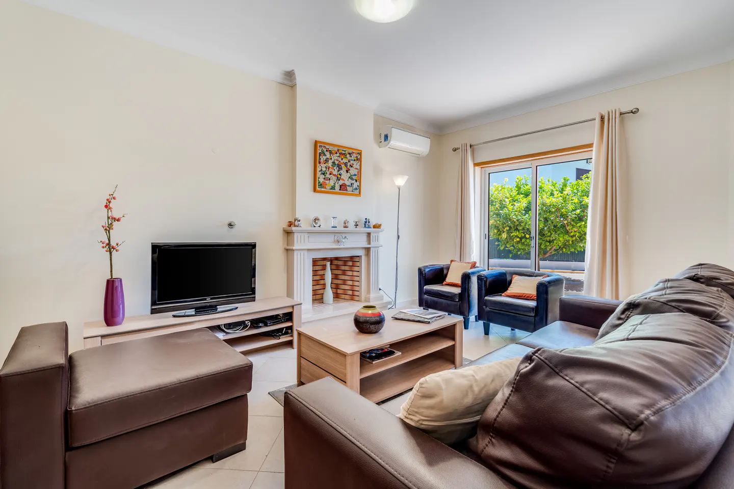Living room with brown leather sofa, two black armchairs, TV, fireplace, and a window with green trees outside.