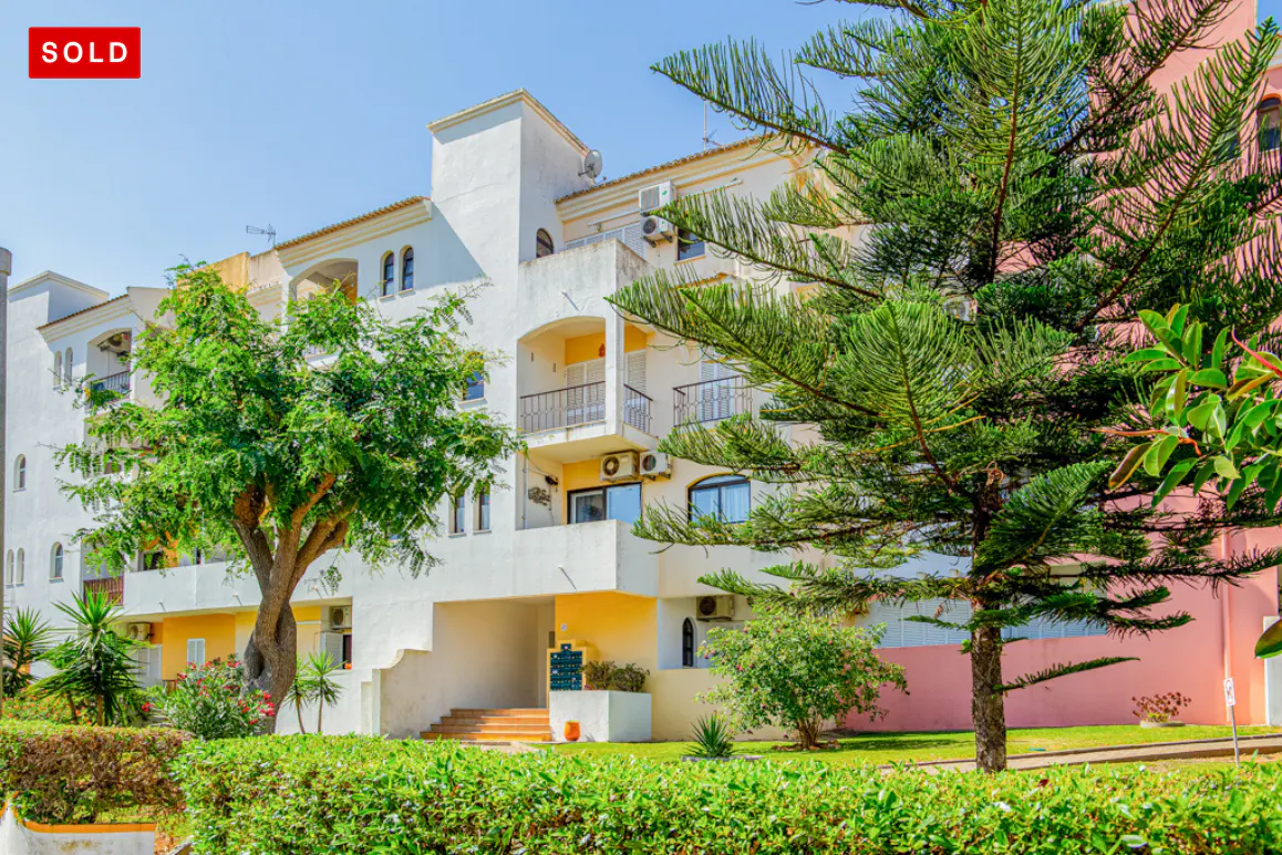 Exterior of a multi-story building with white and yellow walls, balconies, and green trees. A red "SOLD" tag is in the upper left corner.