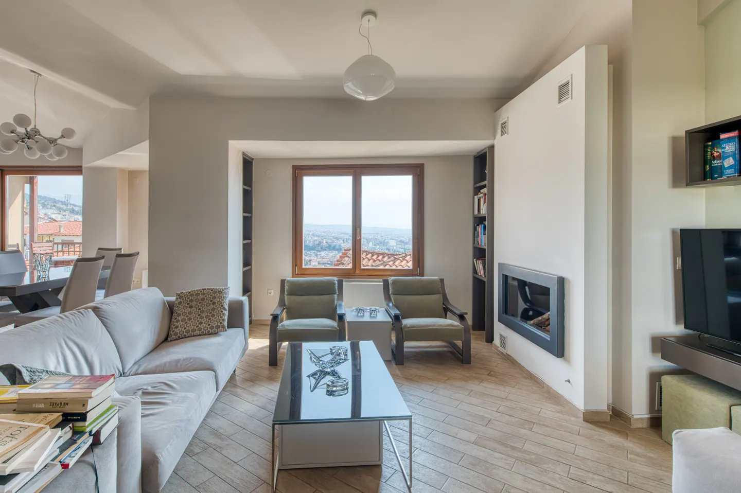 Living room with a gray sofa, two green armchairs, and a glass coffee table. A window overlooks a city view. Fireplace and TV on the right.