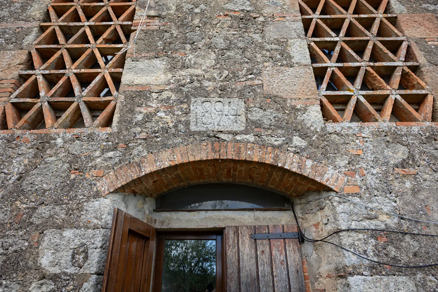 Stone building facade with two lattice windows, a brick arch over a wooden door, and a stone plaque with "1894" carved on it.