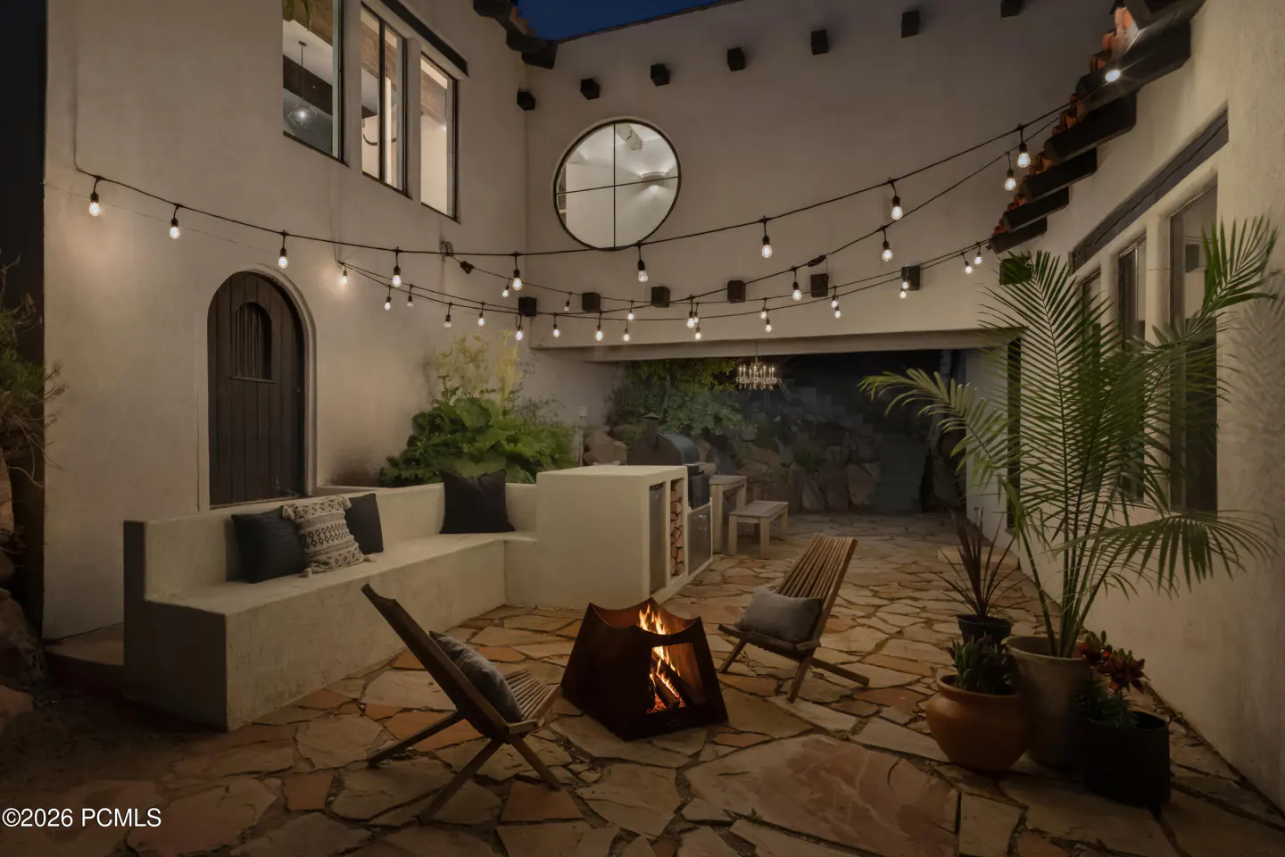 Cozy outdoor patio at dusk with a stone fire pit, built-in bench seating, string lights, and potted plants.