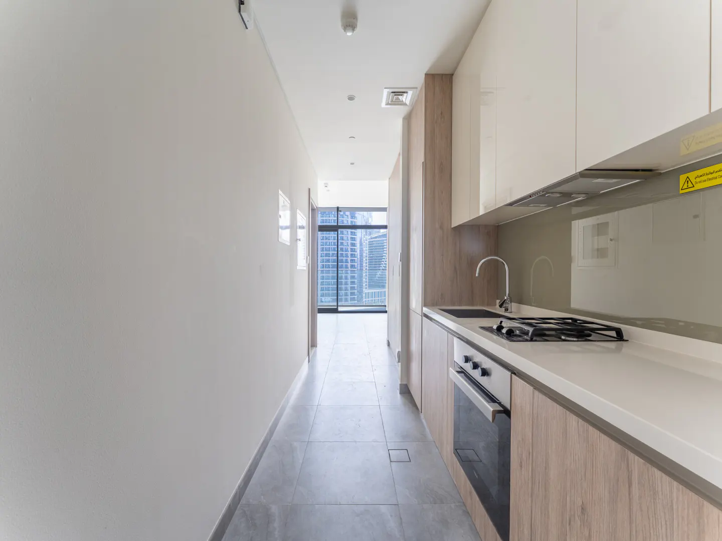 A modern kitchen with white cabinets, a stainless steel sink, and a gas stove. A hallway leads to a window with a city view.
