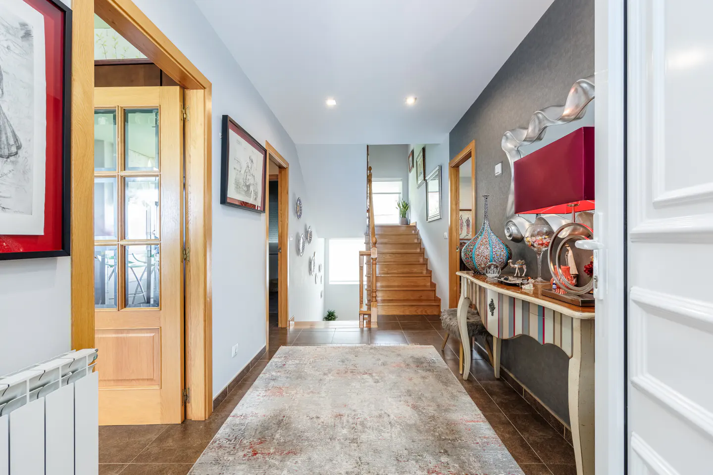 Hallway with brown tile floor, patterned rug, and wooden staircase. A console table with a red lamp and decorative items sits against a gray wall.