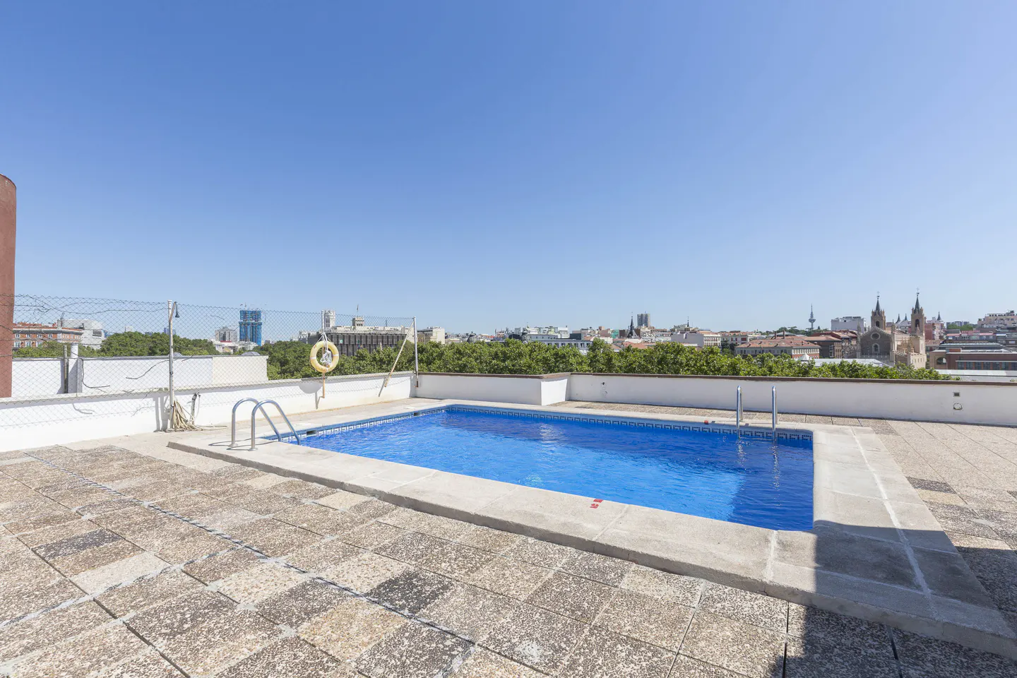 Rooftop pool with blue water and stone deck. A yellow life preserver hangs on a fence with city skyline in the background.