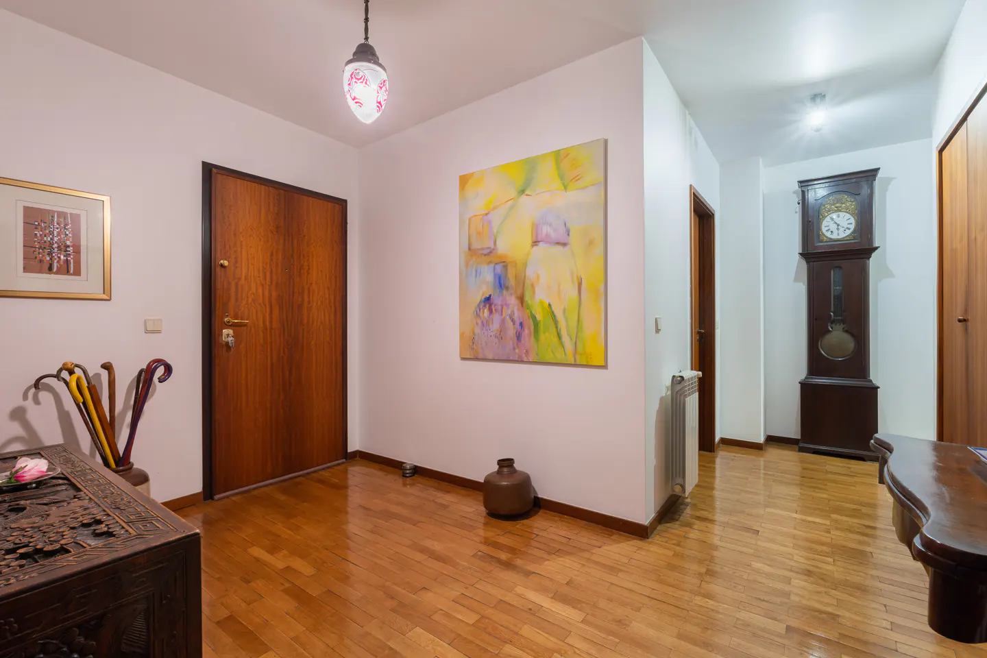 A well-lit foyer with hardwood floors, featuring a wooden door, artwork, and a grandfather clock in the hallway.