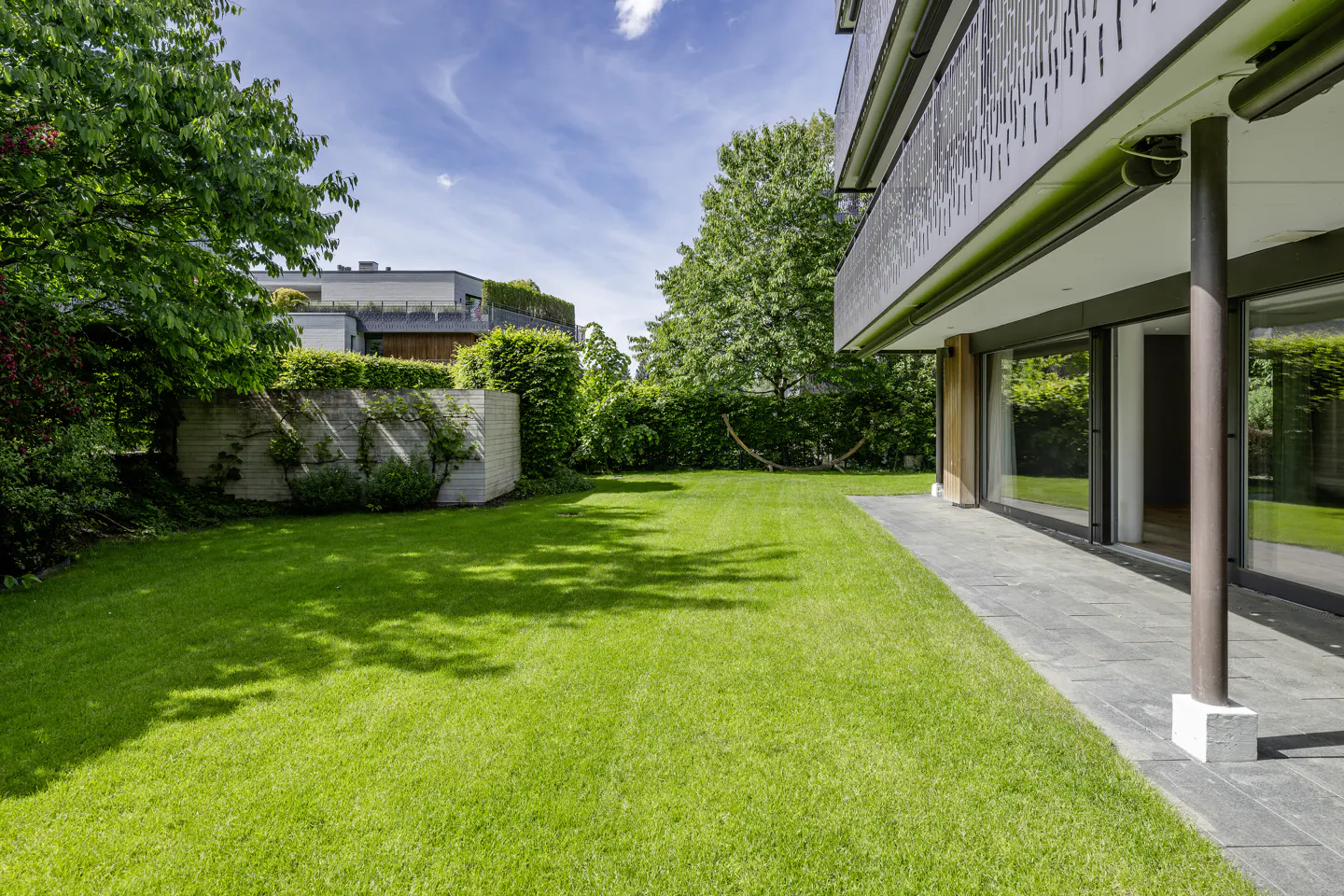 A modern home's backyard with a green lawn, patio, and hammock. The house has large windows and a balcony.