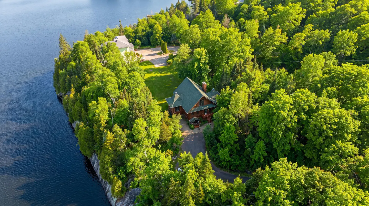 Aerial view of a brown house with a green roof surrounded by lush green trees on a peninsula next to a blue lake.