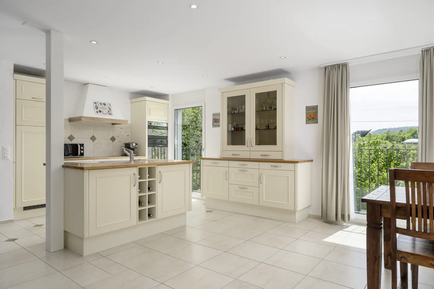 Bright kitchen with cream cabinets, wood countertops, and tile floor. A dining table sits near a window with a view of green trees.