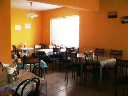 A restaurant dining area with orange walls, tables with patterned tablecloths, and dark wood chairs. A window provides natural light.