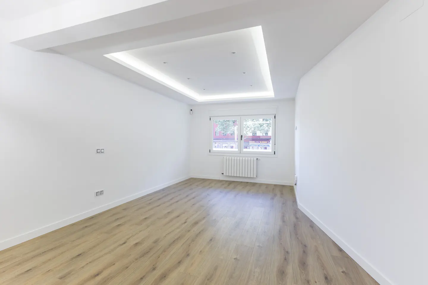 Empty room with white walls, light wood flooring, a window with a radiator, and a modern recessed ceiling light.