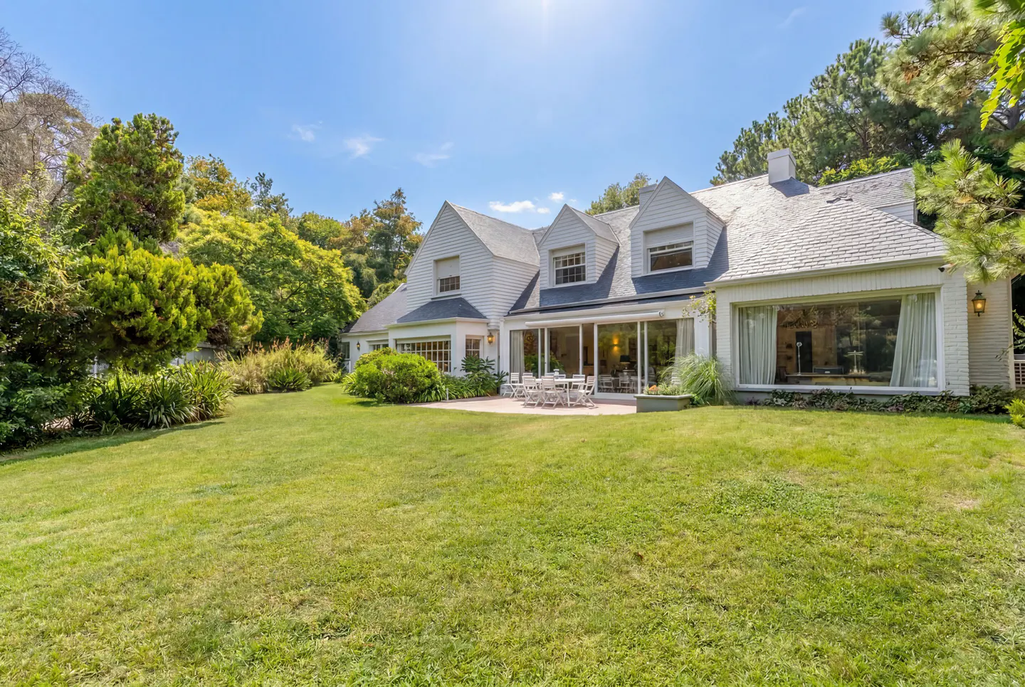 White two-story house with a large lawn, patio furniture, and lush green trees under a clear blue sky.
