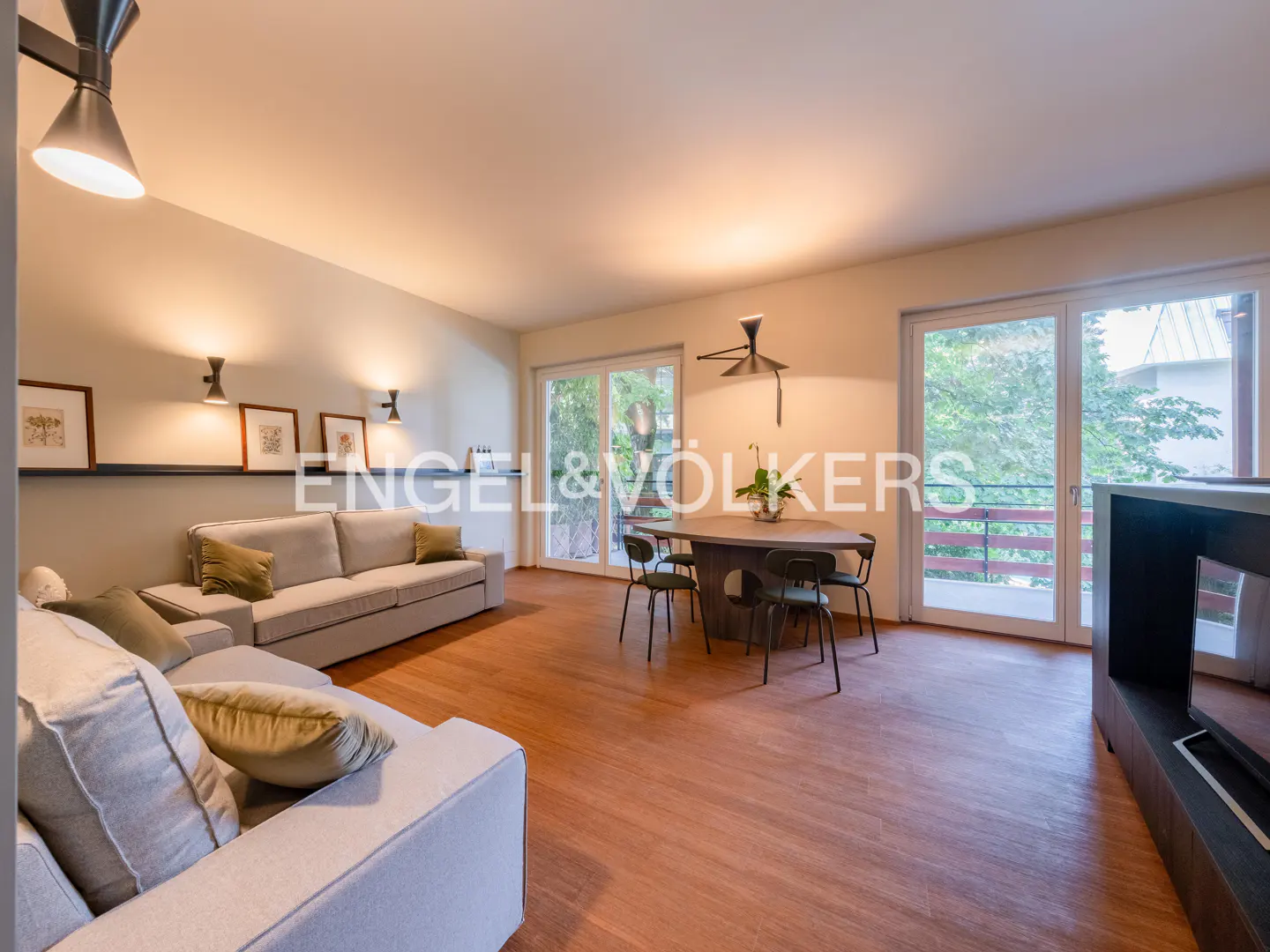 A bright living room with light gray sofas, a round wooden table with four chairs, and wood floors. Balcony doors open to greenery.