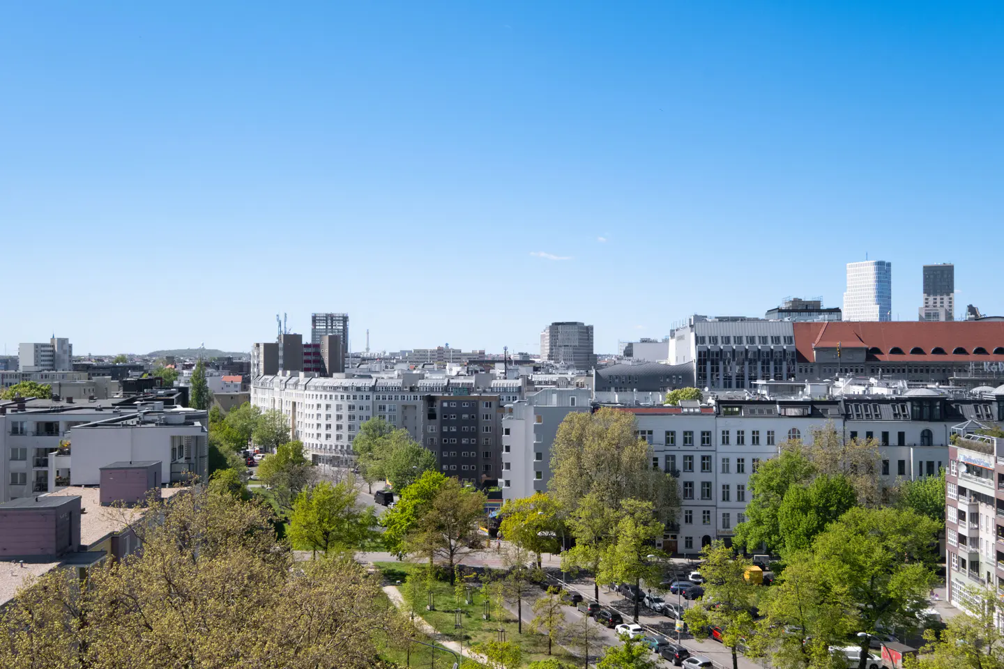 Cityscape view with white buildings, green trees, and a clear blue sky. Cars line the streets below.