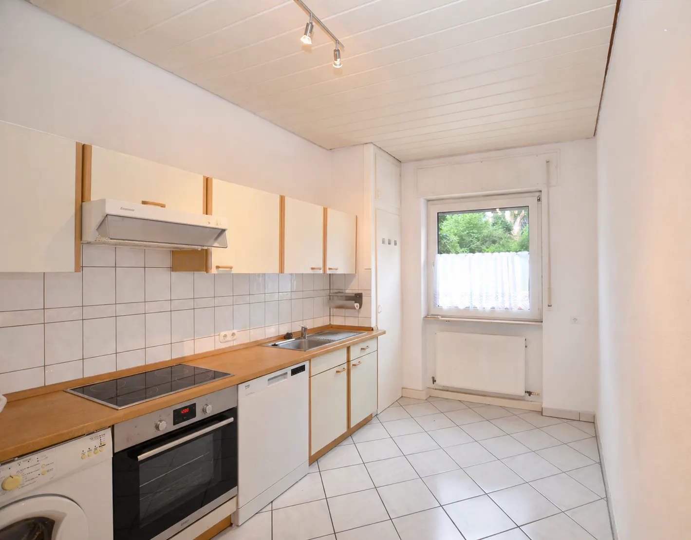 Bright kitchen with white cabinets, wood countertops, and white tiled floors. A window with white curtains lets in natural light.