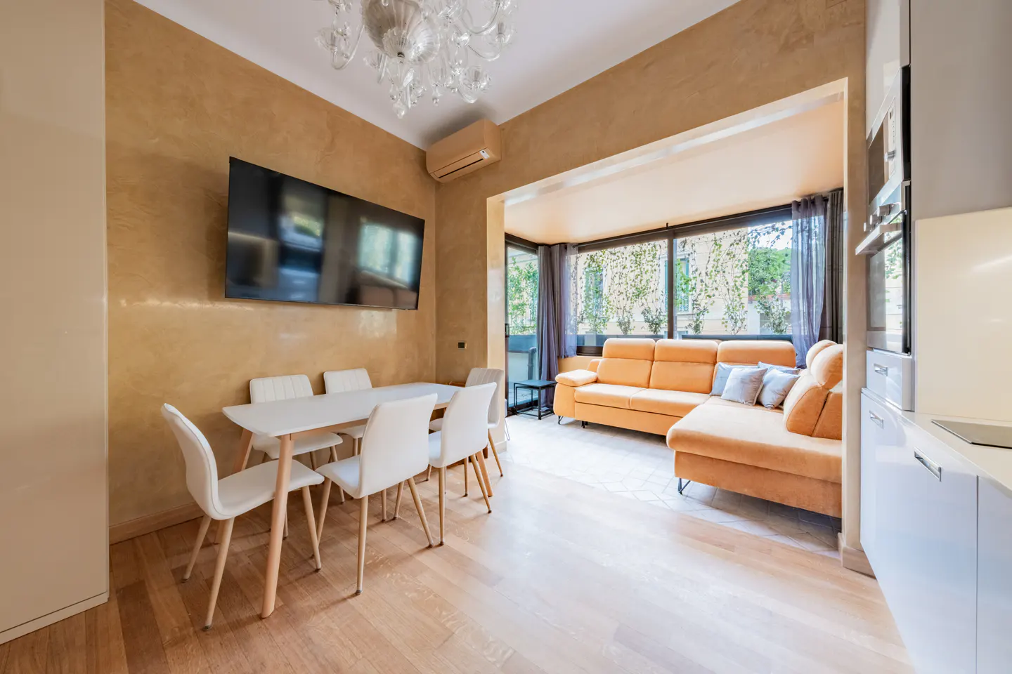 A bright living space with a white table and chairs, a large TV, and an orange sofa near a window.