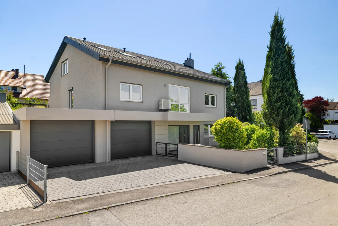 Modern two-story gray house with a double garage, paved driveway, and landscaped garden under a clear blue sky.