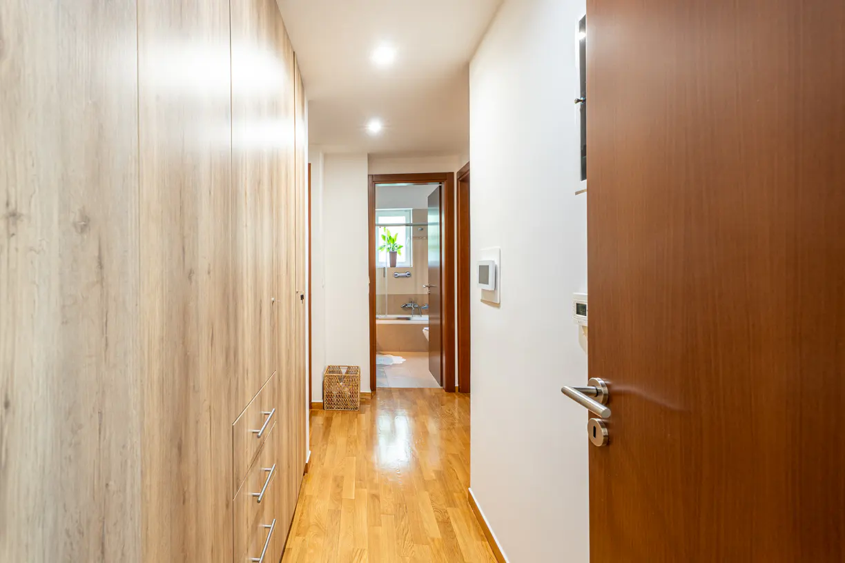 Hallway with wood floors, a light wood closet, and a bathroom visible at the end. A brown door is on the right.