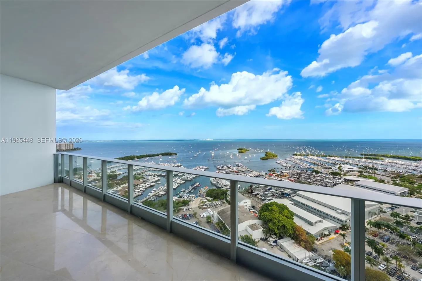Balcony view of a marina filled with boats, under a blue sky with white clouds. Glass railings and a beige floor are visible.