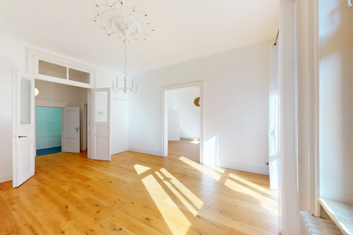 Bright, empty room with wood floors, white walls, and a chandelier. Sunlight streams in through a window with sheer white curtains.