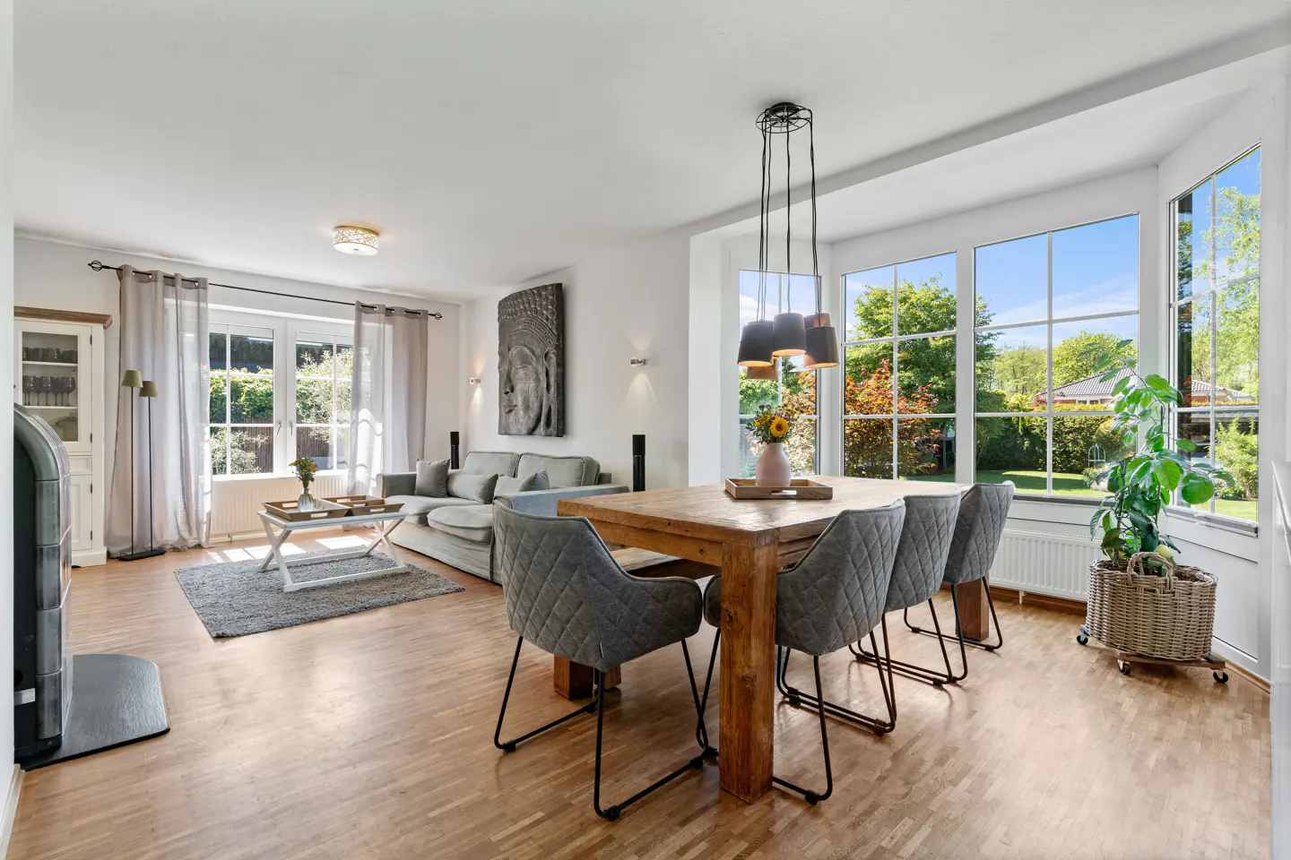 Bright open-plan living and dining room with wooden floors, grey upholstered chairs, and large windows overlooking a garden.