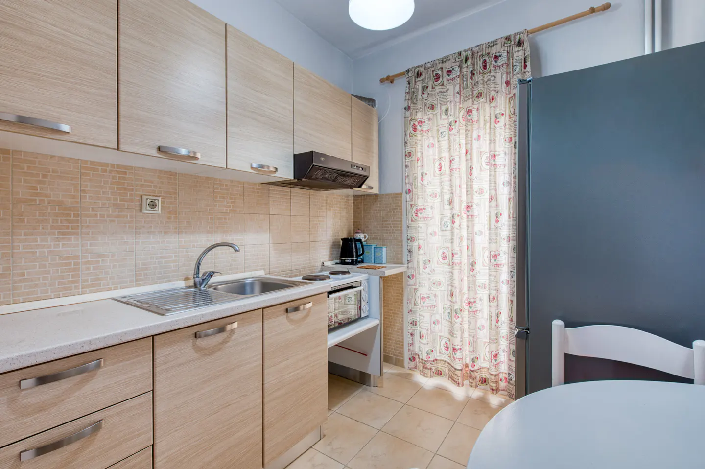 A compact kitchen with light wood cabinets, beige tile backsplash, and a stainless steel sink. A floral curtain hangs near a gray refrigerator.