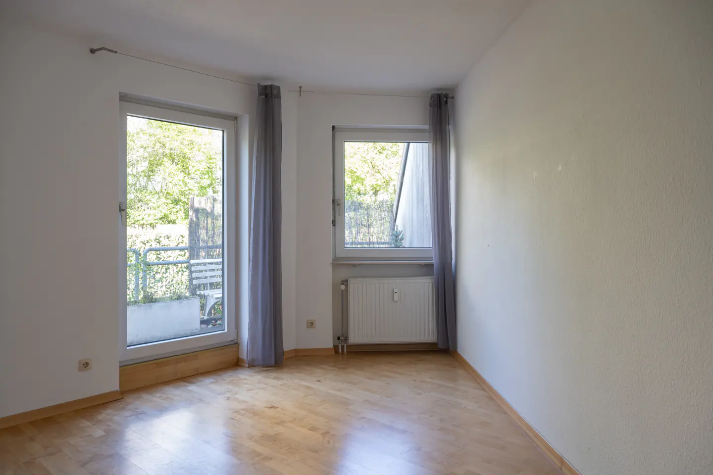 Bright, empty room with wood floors, white walls, and two windows with gray curtains. Balcony visible through the left window.