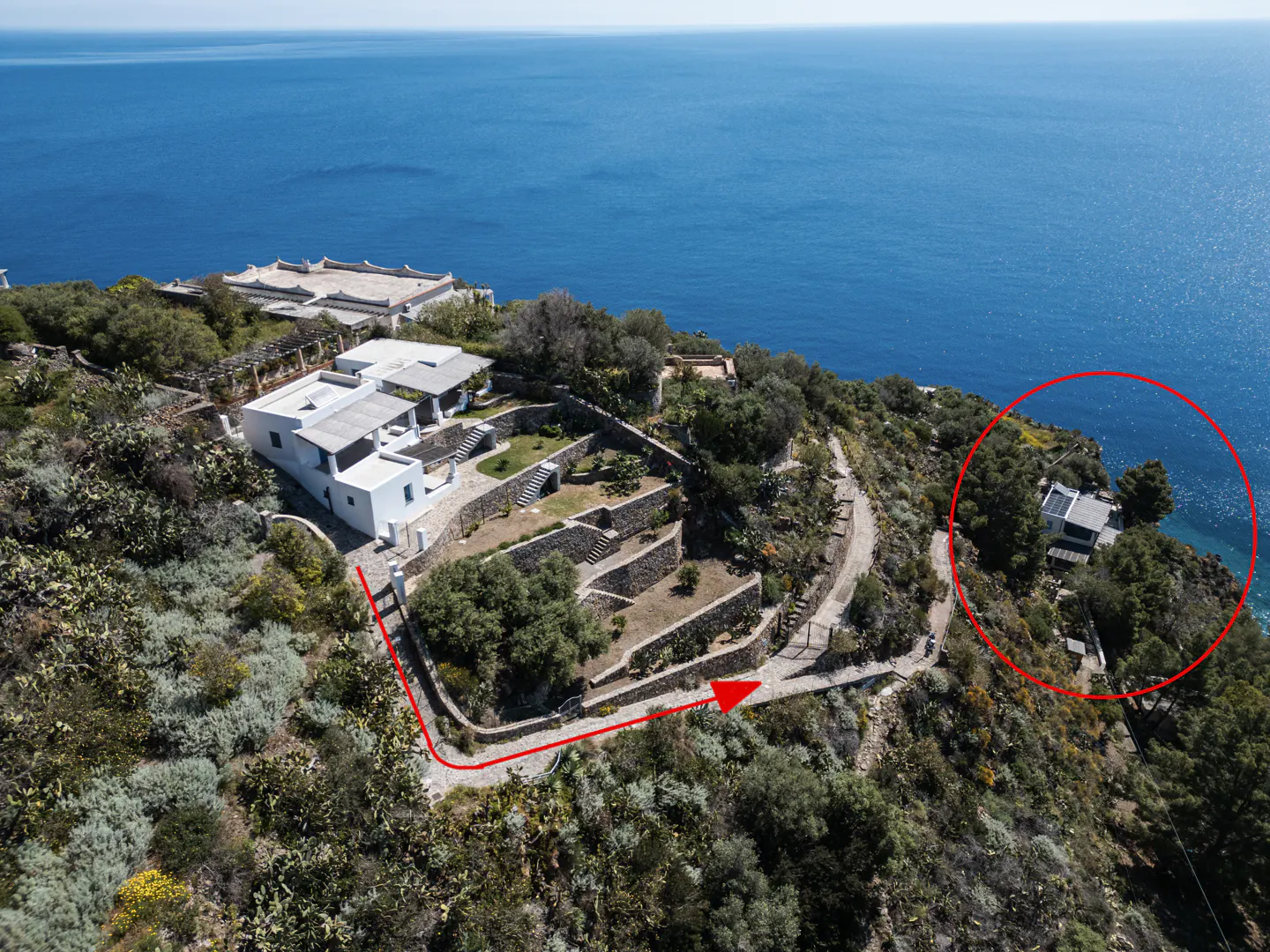 Aerial view of a white villa on a cliffside overlooking the blue sea. A red line indicates a path to another house.