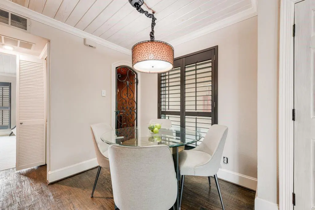 A dining area with a glass table, four light gray chairs, and a decorative light fixture. A window with dark shutters is visible.