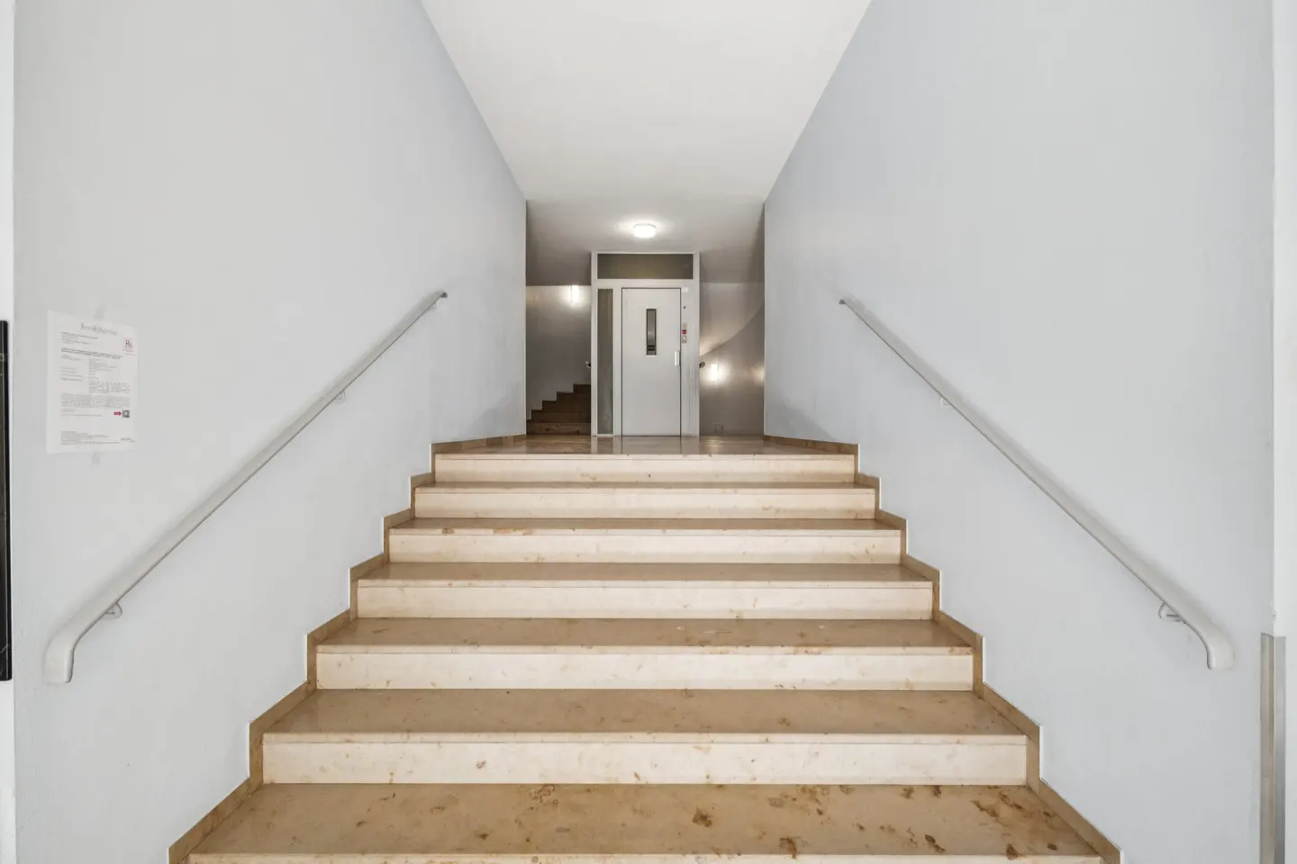 Interior view of marble stairs with metal handrails leading to a white elevator in a white-walled building.
