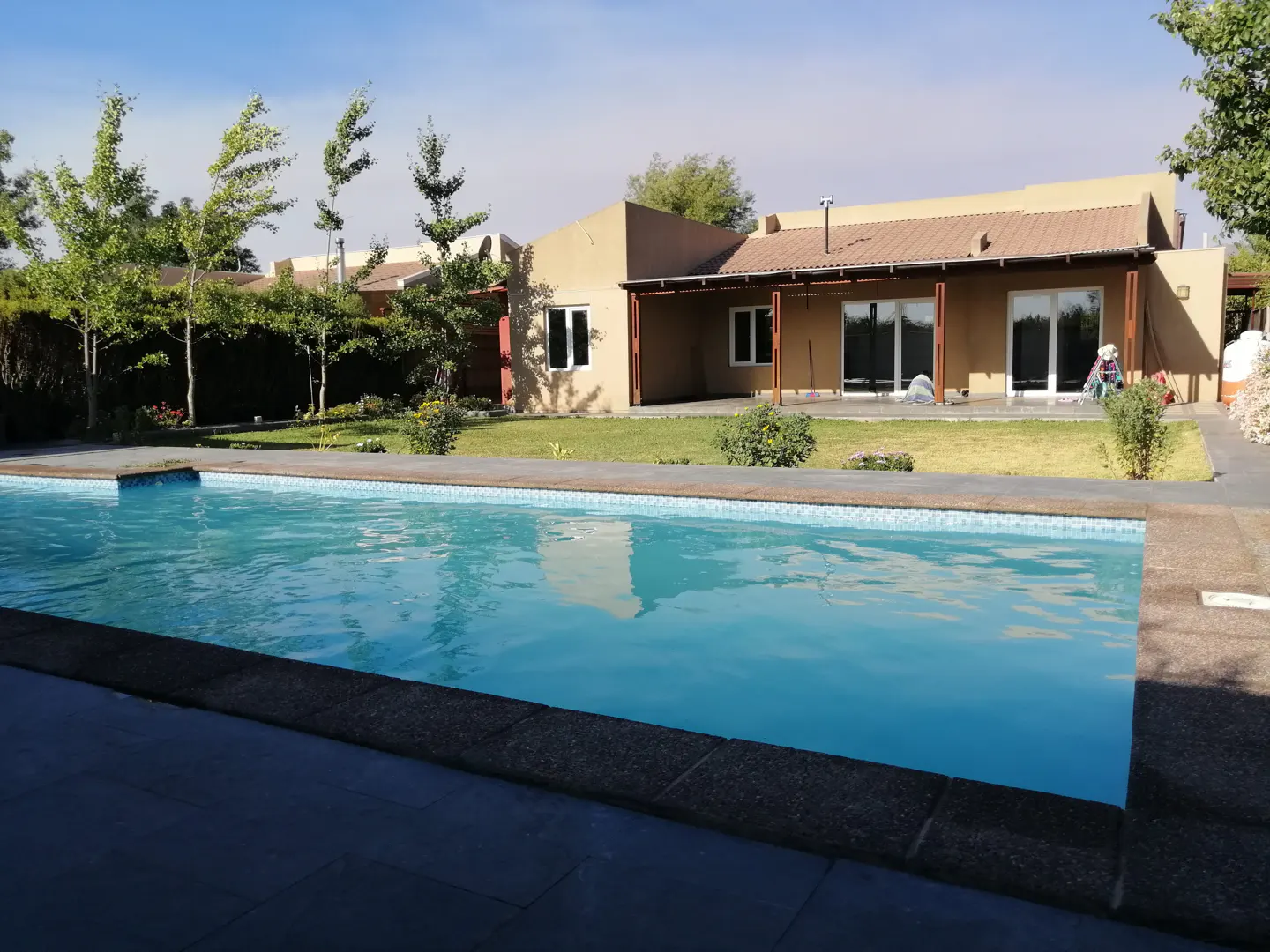 A tan house with a brown roof overlooks a blue swimming pool on a sunny day. Trees and green grass surround the house.