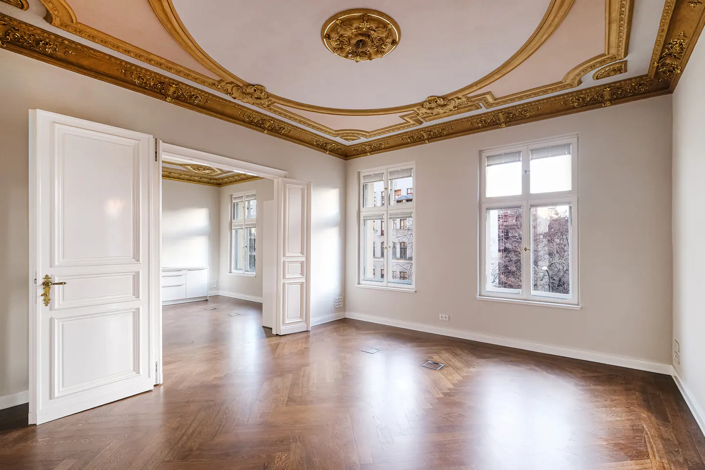Bright, empty room with herringbone wood floors, white walls, and ornate gold ceiling trim. White doors lead to another room. Two windows show a city view.