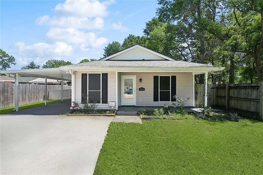 A single-story beige house with black shutters, a white door, and a carport. Green lawn and trees surround the property.