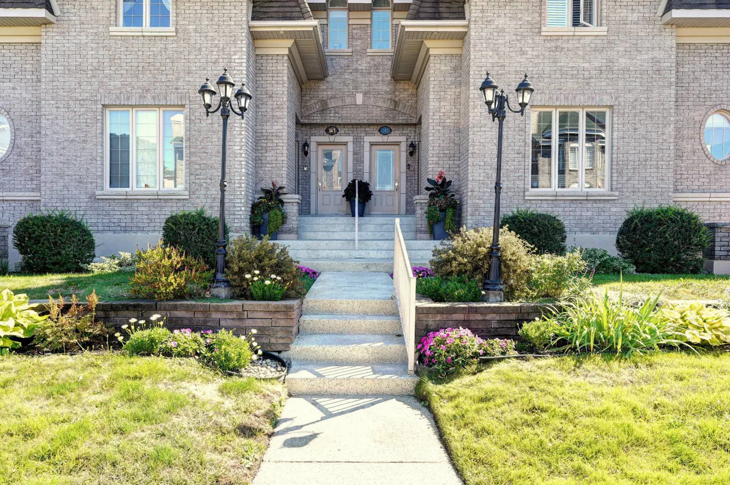 Exterior view of a two-story brick apartment building with a concrete walkway leading to the entrance. Black lampposts flank the stairs and colorful flowers line the walkway.