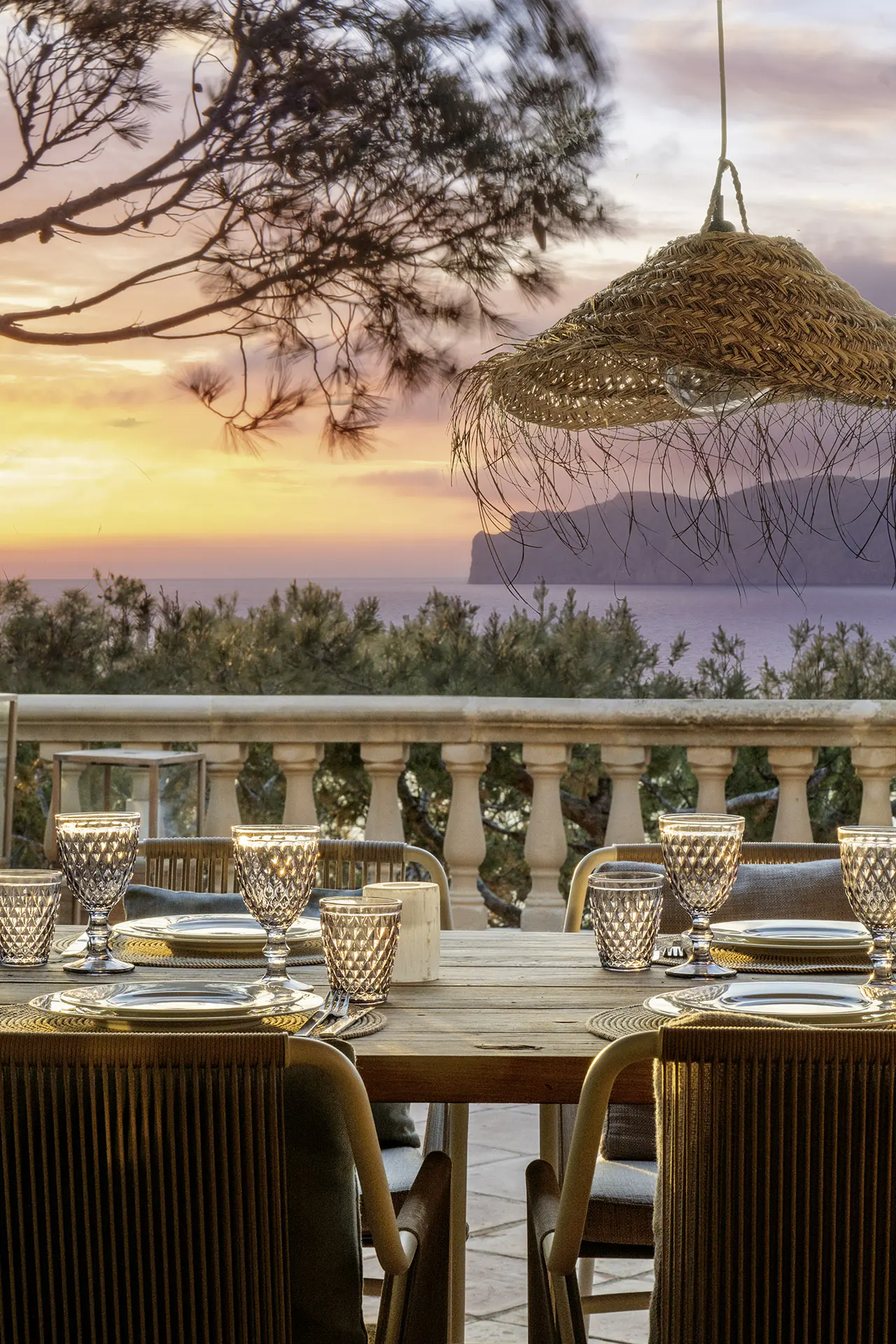Outdoor dining area with a wooden table set for a meal, overlooking the ocean at sunset. A straw lampshade hangs above.