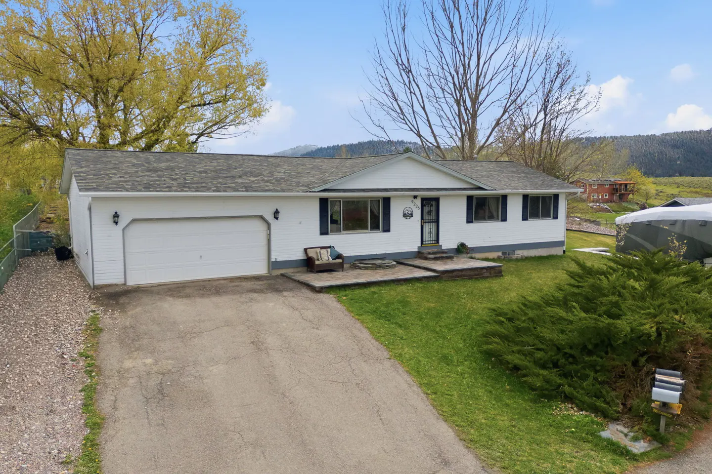 A white, single-story house with a gray roof, blue shutters, and an attached garage. A driveway leads to the house, and a green lawn surrounds it.