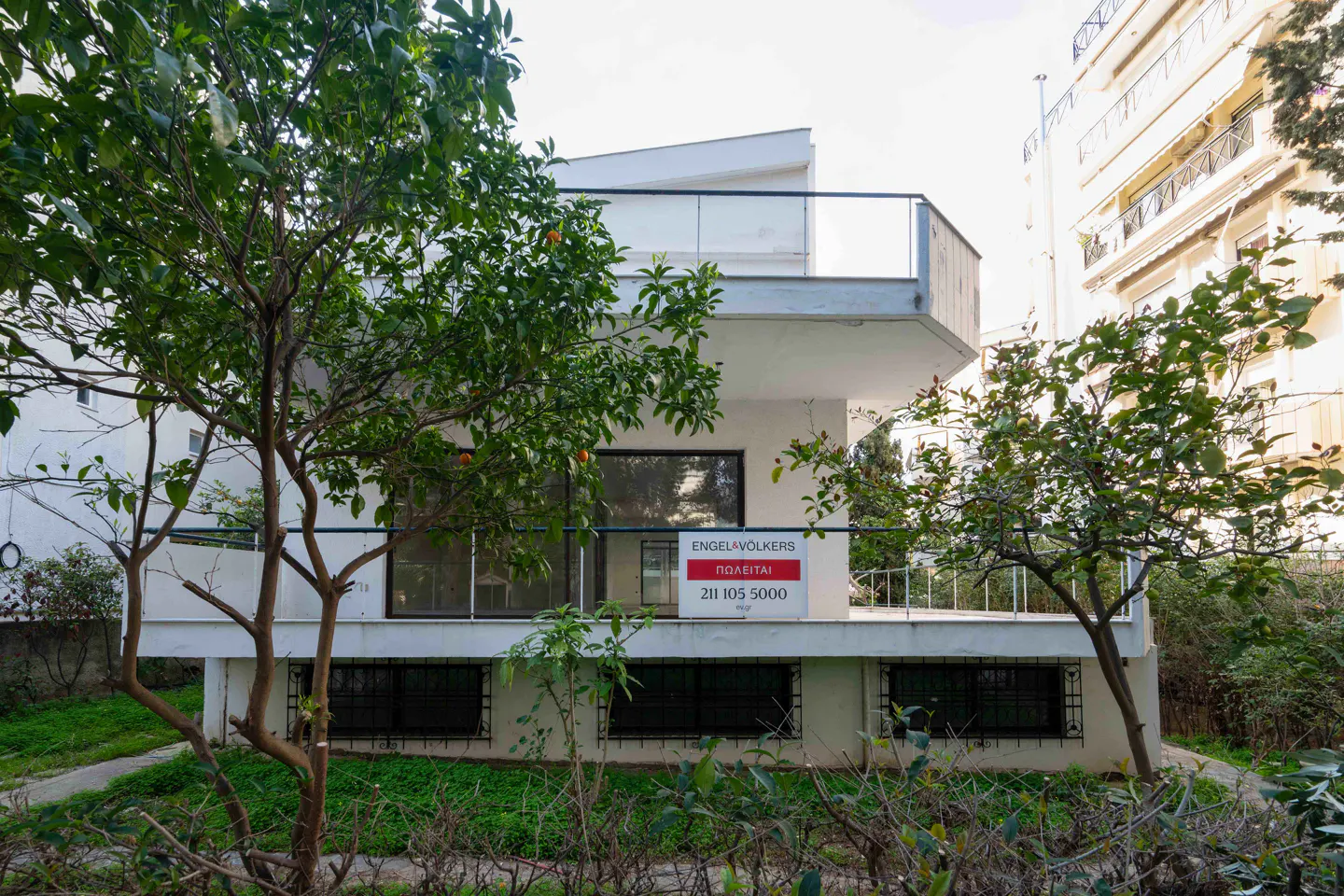 Two-story white house with a balcony and an Engel & Völkers "For Sale" sign, partially obscured by trees.