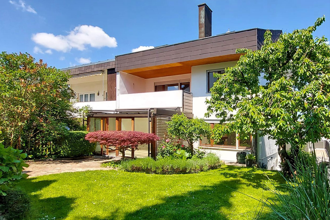 Modern white house with a brown roof, balcony, glass-enclosed patio, and a lush green garden with a red Japanese maple.