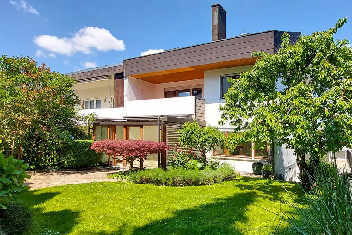 Modern white house with a brown roof, balcony, glass-enclosed patio, and a lush green garden with a red Japanese maple.