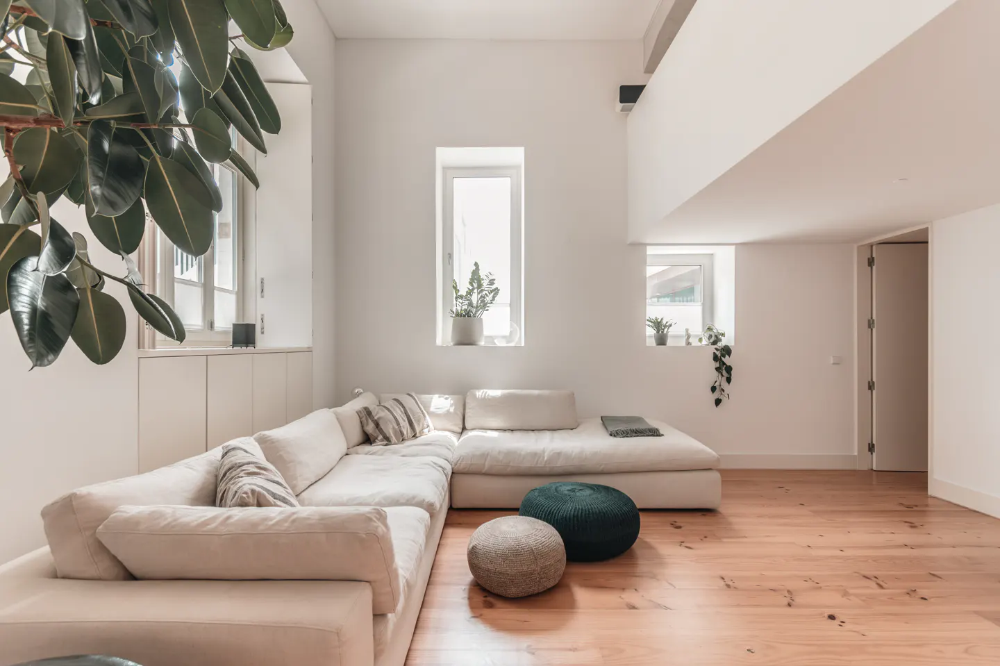 Bright living room with a large white sectional sofa, wooden floors, and two poufs. A large plant is in the corner.