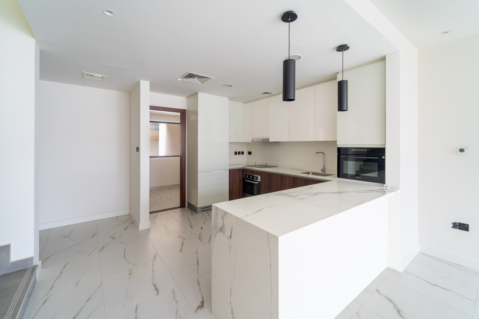 Bright, modern kitchen with white cabinets, marble countertops, and dark wood accents. Two black pendant lights hang above the island. Tiled floor.