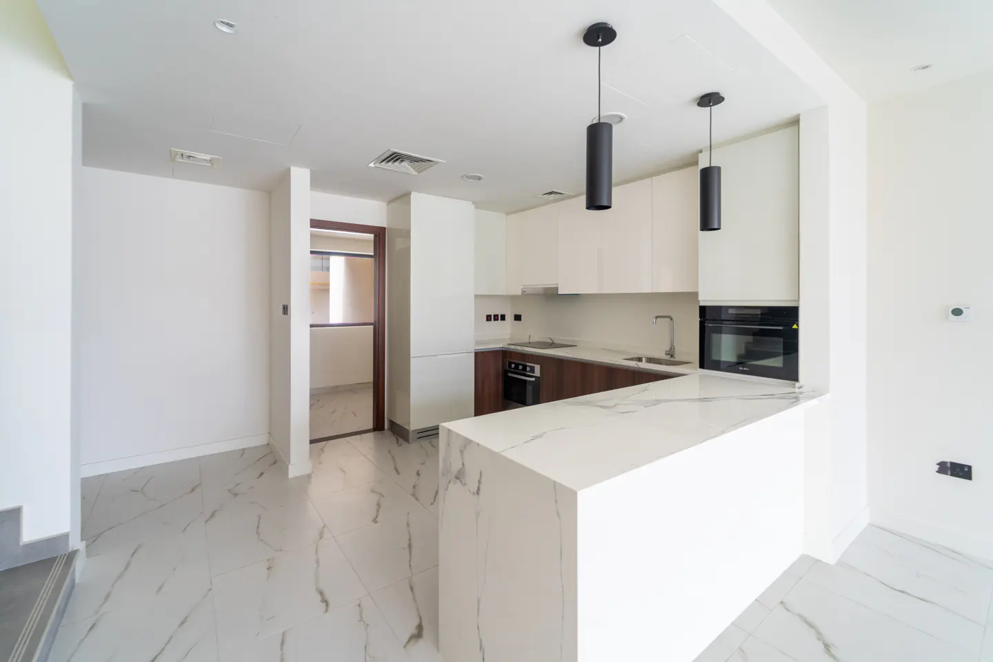 Bright, modern kitchen with white cabinets, marble countertops, and dark wood accents. Two black pendant lights hang above the island. Tiled floor.