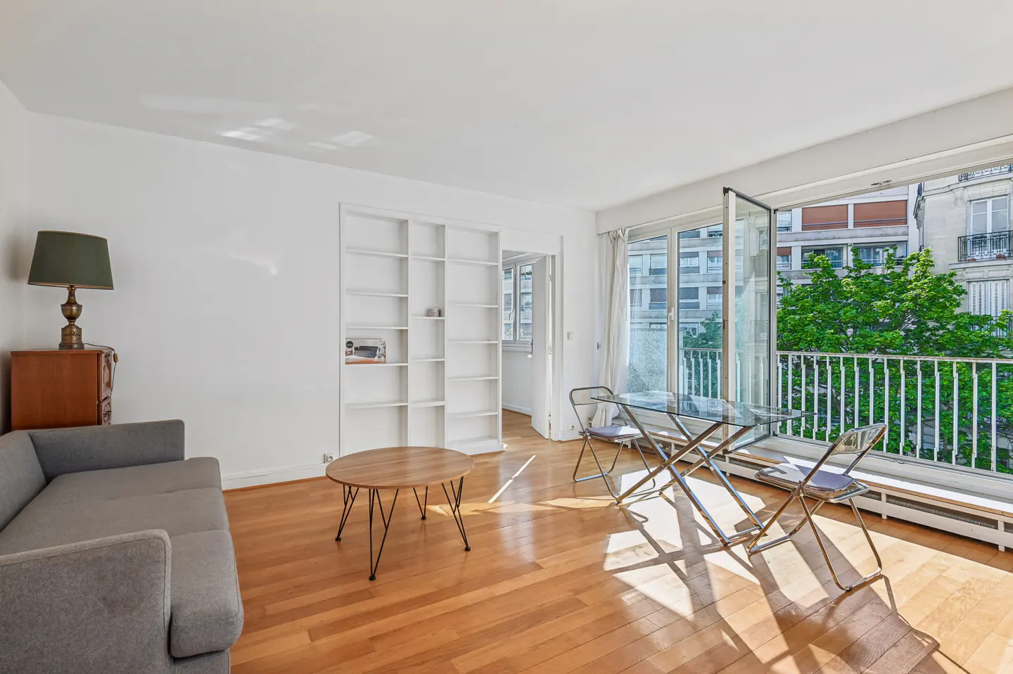 Bright living room with hardwood floors, gray sofa, round wood table, and glass table with chairs near open balcony doors.