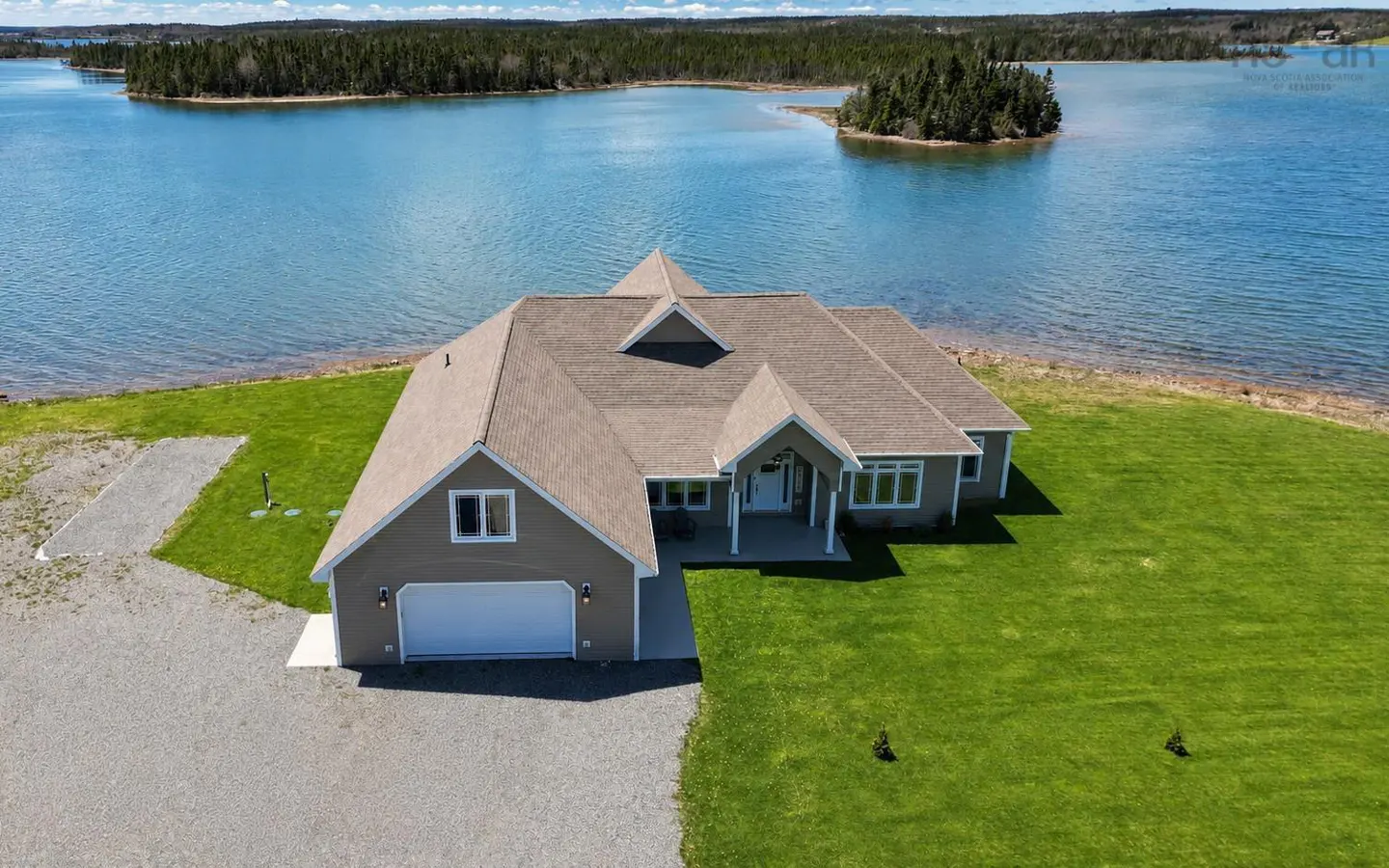 Aerial view of a tan house with a white garage, surrounded by a green lawn and a scenic blue bay with forested islands.