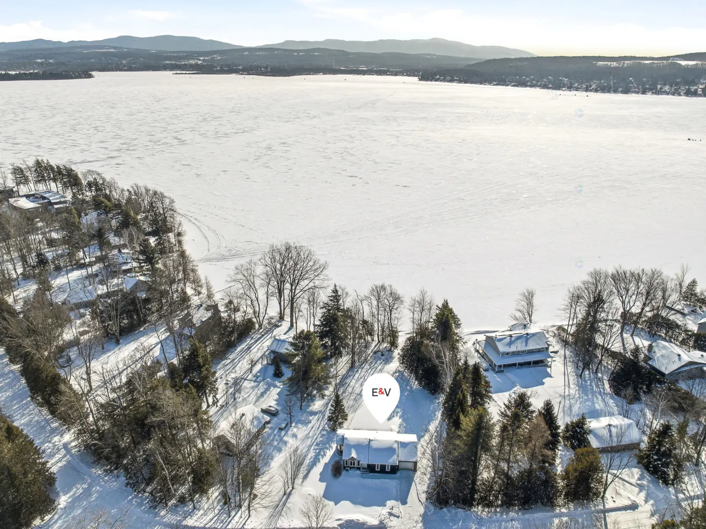 Aerial view of a snow-covered house near a frozen lake, surrounded by trees. Mountains are visible in the background under a clear sky.