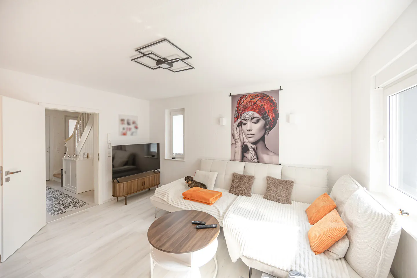 Bright living room with white walls, light wood floors, and a white sectional sofa. A large portrait hangs above the sofa. A round coffee table sits in front.