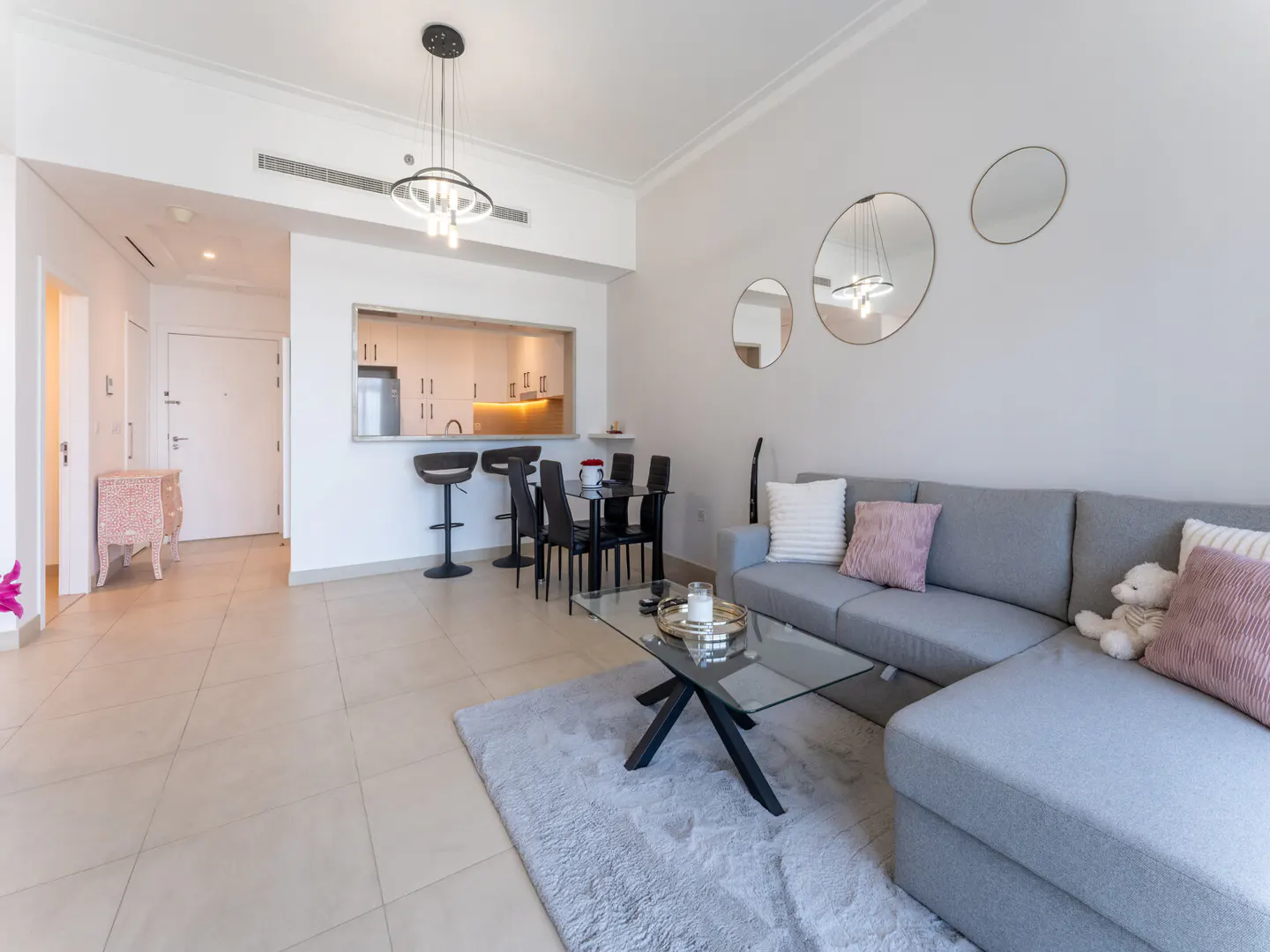 A bright, modern living space with a gray sectional sofa, glass coffee table, and open kitchen with bar seating.