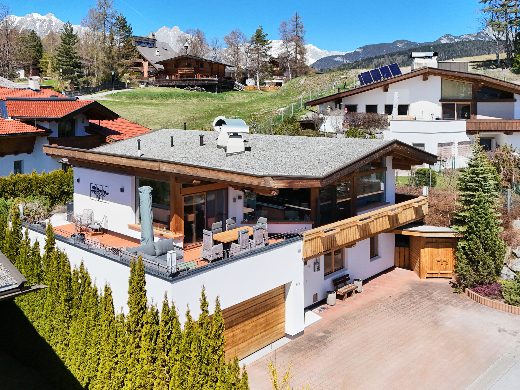 Modern white house with a wooden garage, balcony dining area, and mountain views in a sunny, scenic neighborhood.
