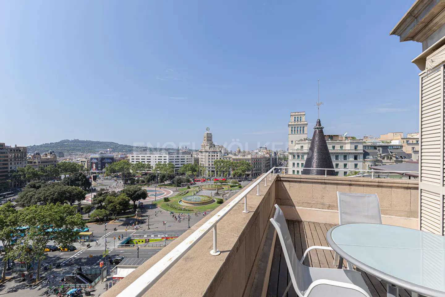 Balcony view of Plaça de Catalunya in Barcelona. Table and chairs on a wooden deck overlook the city square with buildings and a blue sky.